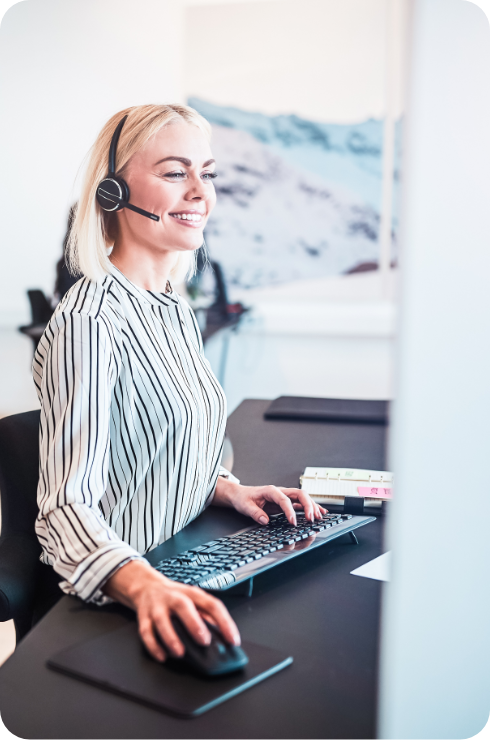karriere tutor Eine blonde Frau lächelt und arbeitet an einem Schreibtisch. Sie trägt ein schwarz-weiß gestreiftes Hemd, ein Headset und tippt mit einer Maus auf einer Tastatur. Im Hintergrund ist eine verschwommene, verschneite Landschaft zu sehen.