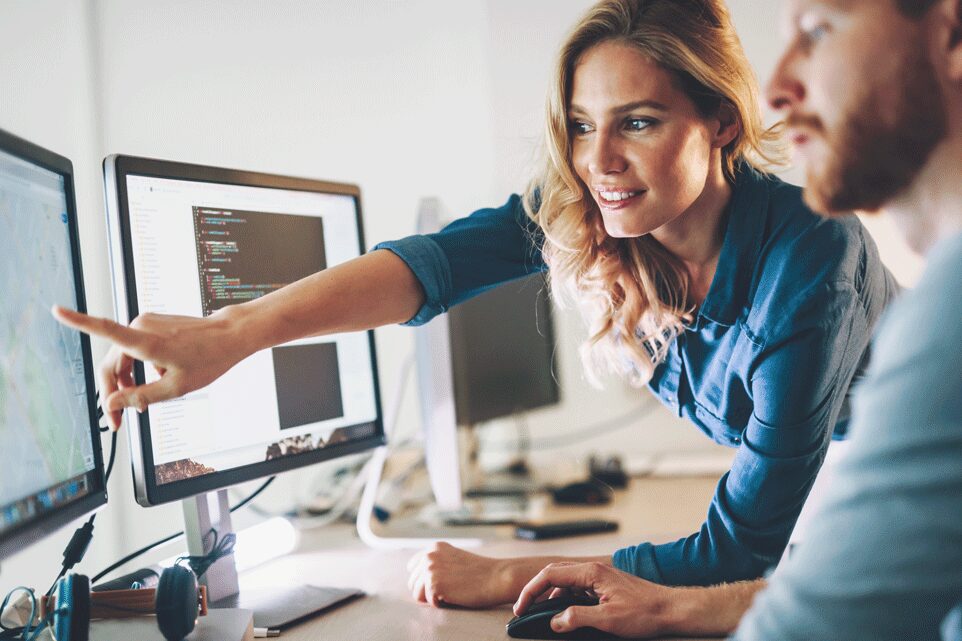 karriere tutor Eine Frau mit blonden Haaren und blauem Hemd zeigt auf einen Computerbildschirm, während sie einem bärtigen Mann neben ihr etwas erklärt. Beide konzentrieren sich auf die Monitore, auf denen Code und eine Karte angezeigt werden. Sie scheinen in einem Büro zu arbeiten.