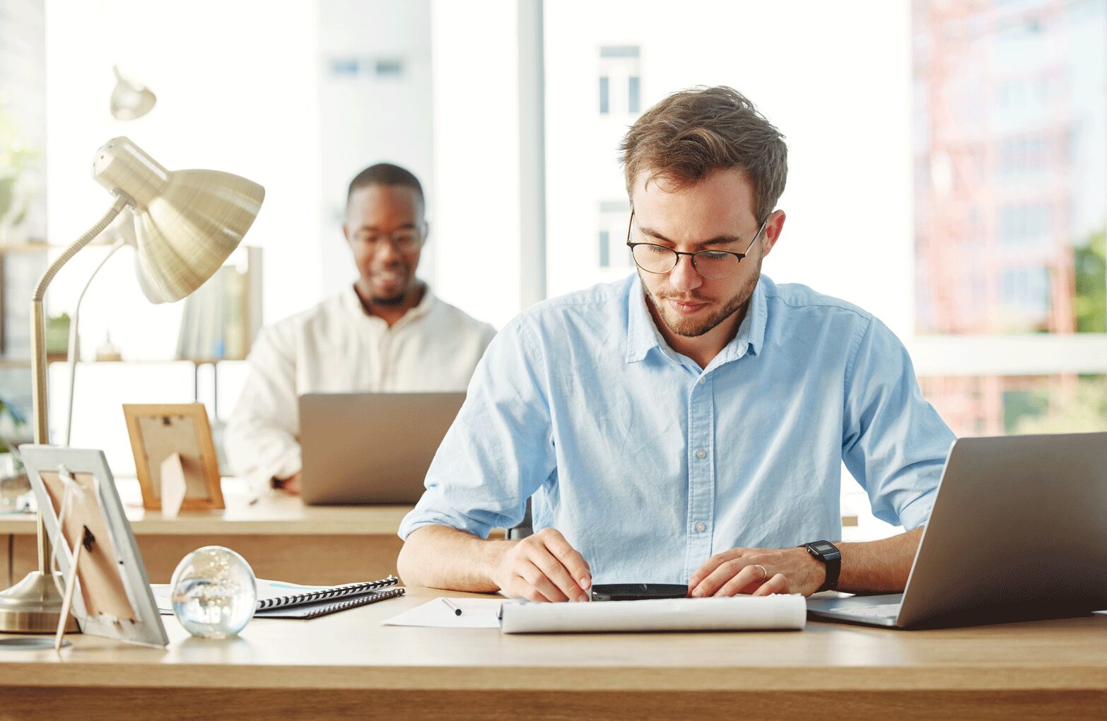 karriere tutor Zwei Männer arbeiten an Schreibtischen in einem hell erleuchteten Büro. Der Mann im Vordergrund, der eine Brille und ein hellblaues Hemd trägt, ist konzentriert darauf, in ein Notizbuch zu schreiben, neben ihm steht ein Laptop. Im Hintergrund arbeitet ein anderer Mann an einem Laptop und lächelt.