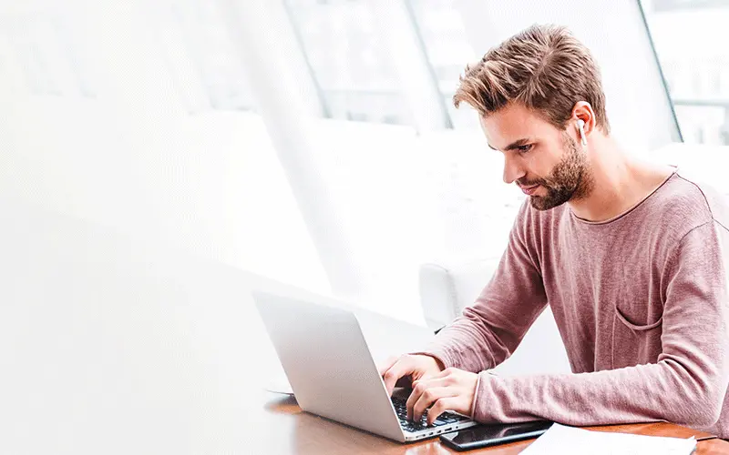 karriere tutor® Ein Mann mit Bart und rosa Hemd tippt an einem Schreibtisch in einem hell erleuchteten Raum auf einem Laptop.