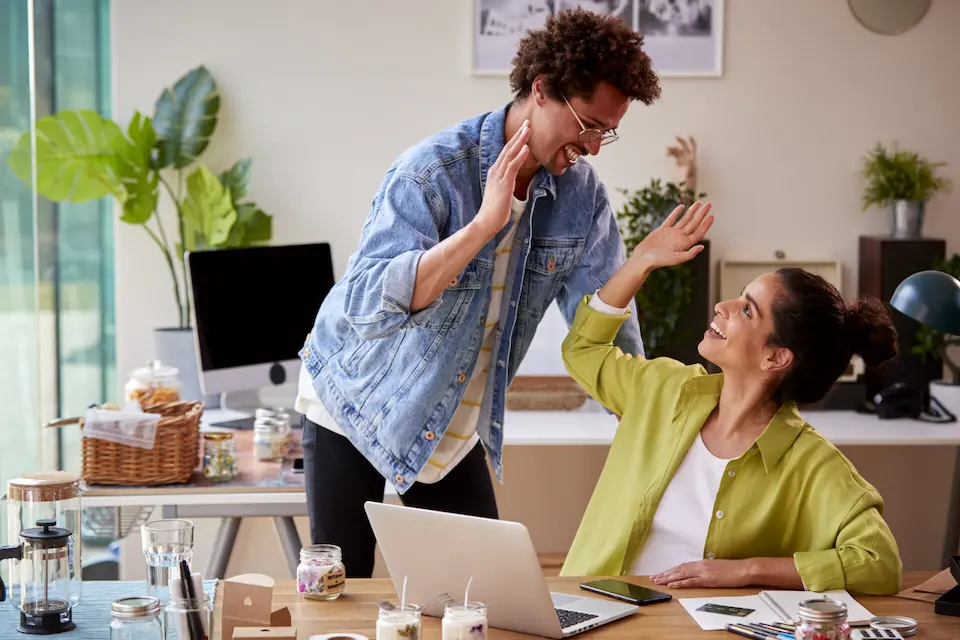 karriere tutor® Zwei Personen in einem Home-Office geben sich High Fives und feiern Erfolge. Auf dem Schreibtisch stehen ein Laptop und Kerzen.