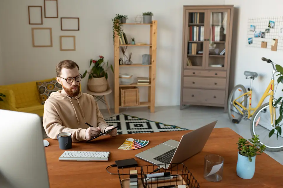 karriere tutor® Eine Person arbeitet in einem Heimbüro an einem Schreibtisch, umgeben von einem Laptop, einer Tastatur und Farbmustern, an einem Tablet.