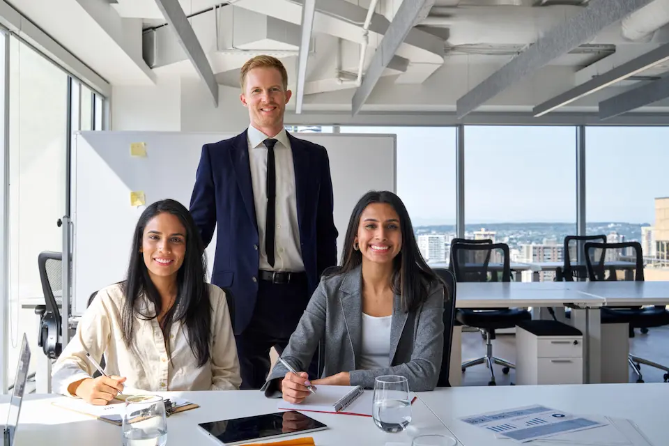 karriere tutor® Drei Personen in einem modernen Büro: zwei Frauen sitzen mit Notizbüchern an einem Schreibtisch und ein Mann steht hinter ihnen, alle lächeln.