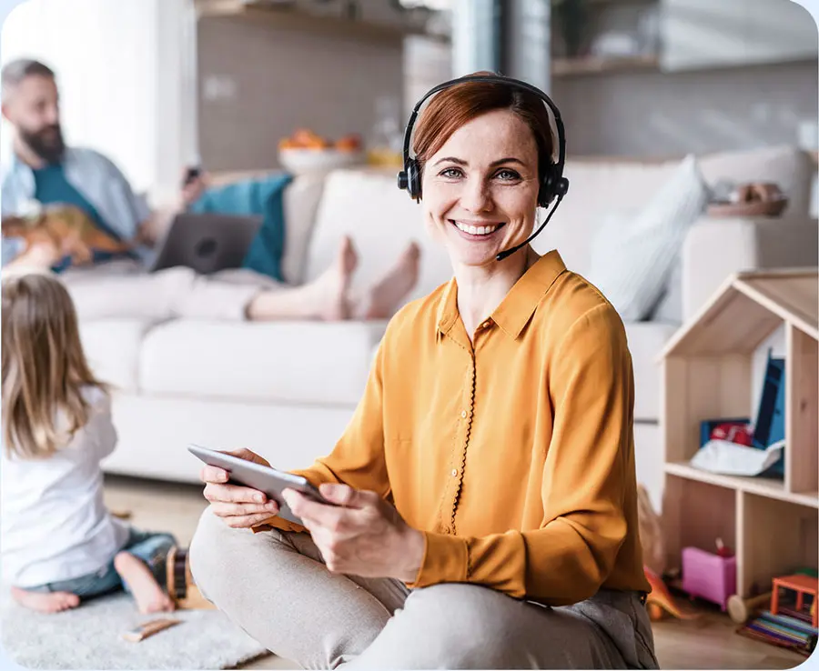 karriere tutor® Eine Frau mit Headset, die wahrscheinlich gerade ihre Weiterbildung zum Personalfachkaufmann absolviert, lächelt und hält ein Tablet in der Hand. In der Nähe spielt ein Kind auf dem Boden und im Hintergrund arbeitet ein Mann an seinem Laptop.