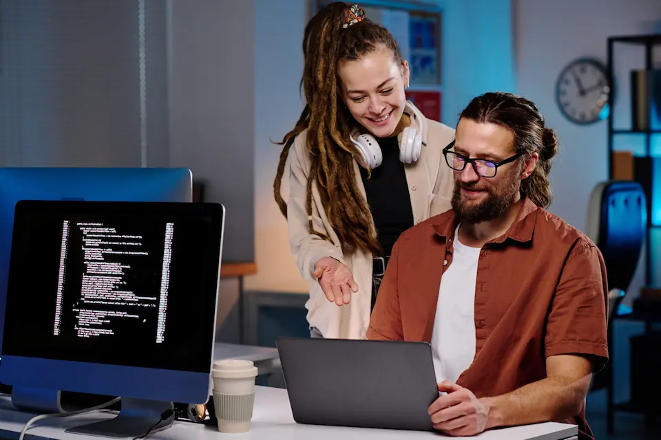 karriere tutor® Zwei Personen lächeln, während sie an einem Schreibtisch in einem Büro an Laptops arbeiten. Auf einem der Computerbildschirme wird Code angezeigt.