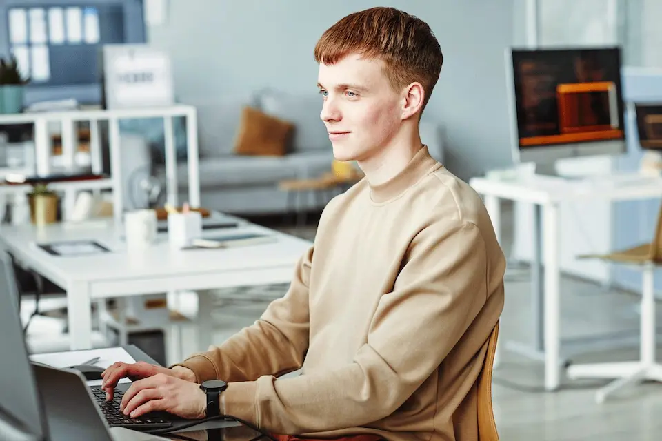 karriere tutor® Junger Mann in einem beigen Sweatshirt arbeitet in einem modernen Büro an einem Laptop.