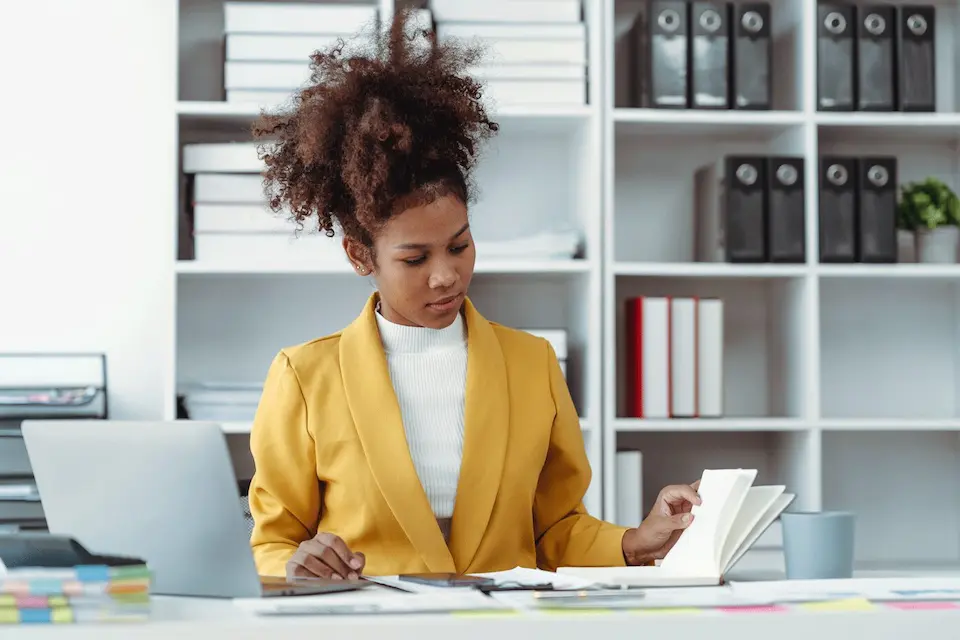 karriere tutor® Eine Frau in einem gelben Blazer sitzt an einem Schreibtisch und liest ein Buch in einem Büro, hinter ihr stehen Regale voller Ordner.