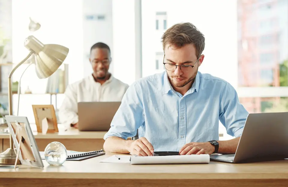 karriere tutor® Zwei Männer in einem Büro. Einer nutzt ein Tablet am Schreibtisch vor ihm, der andere arbeitet im Hintergrund an einem Laptop.