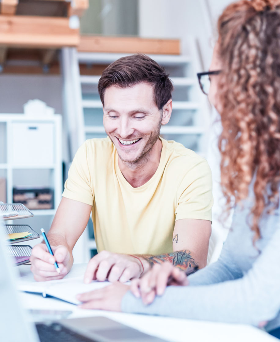 karriere tutor® Zwei Menschen lächeln und arbeiten an einem Schreibtisch in einem hellen Büro mit Regalen und Treppen im Hintergrund.