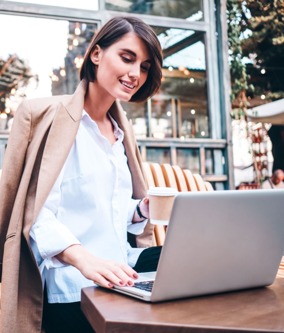 karriere tutor® Frau im hellen Mantel, lächelnd, während sie in einem Straßencafé an einem Laptop arbeitet und eine Kaffeetasse hält.