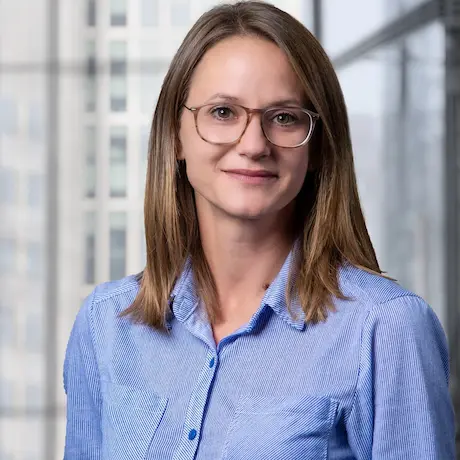 karriere tutor® Eine Frau mit Brille und blauem Hemd lächelt in einem Büroumfeld mit großen Fenstern und Stadtgebäuden im Hintergrund.