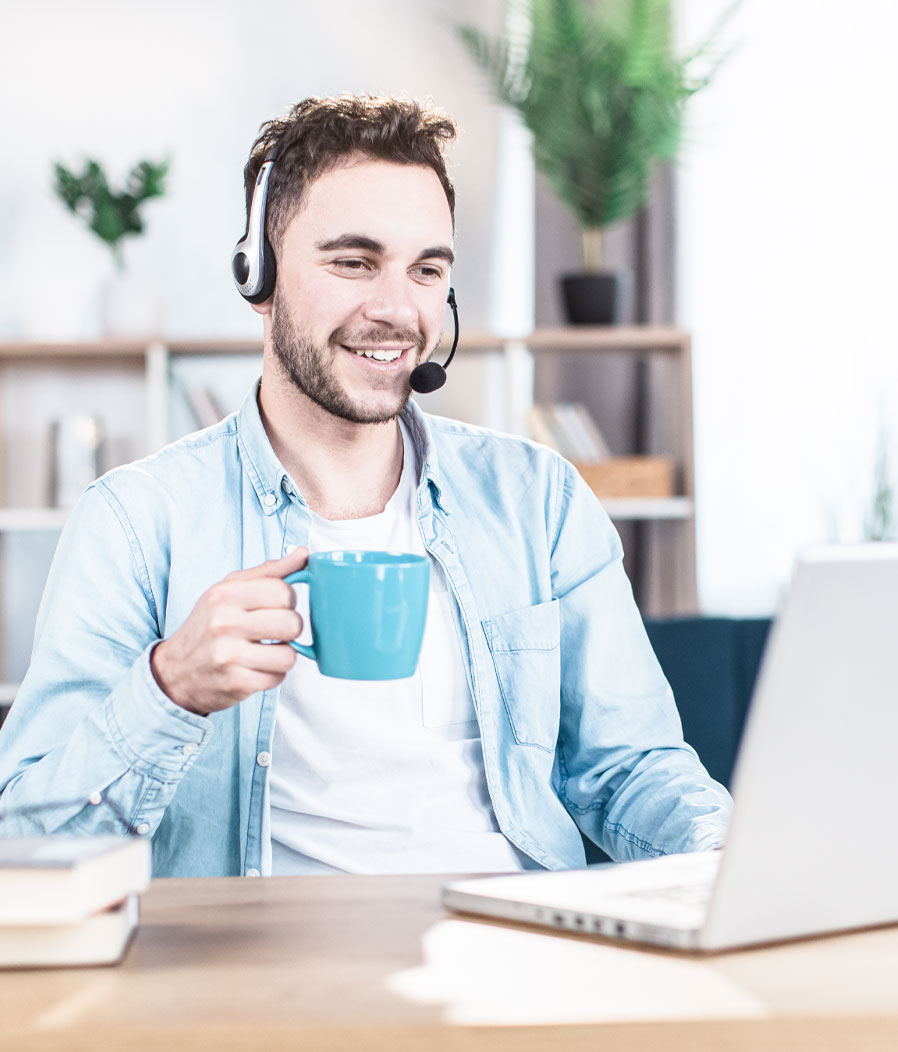 karriere tutor® Ein lächelnder Mann mit Headset hält eine blaue Tasse in der Hand, während er an einem Schreibtisch in einem hellen Raum mit Pflanzen an einem Laptop arbeitet.