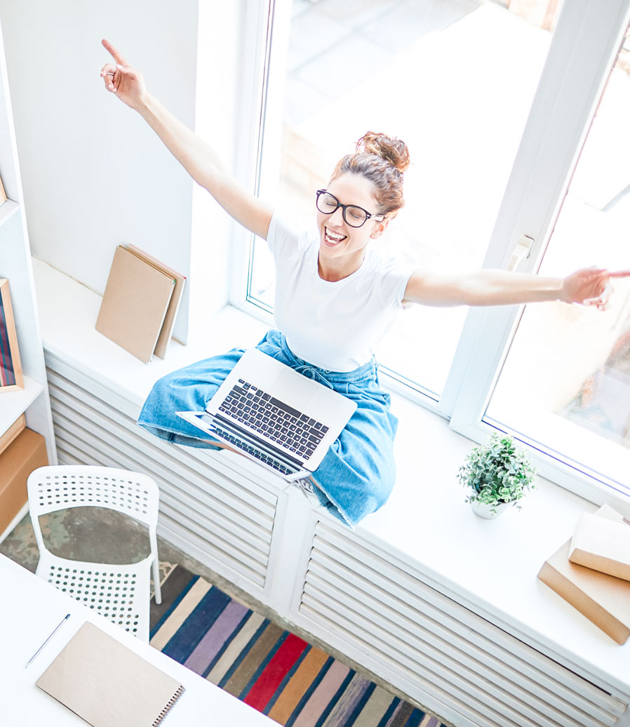 karriere tutor® Person mit Brille und weißem Hemd sitzt mit einem Laptop am Fenster und hat die Arme aufgeregt in die Höhe gehoben, Büroumgebung.