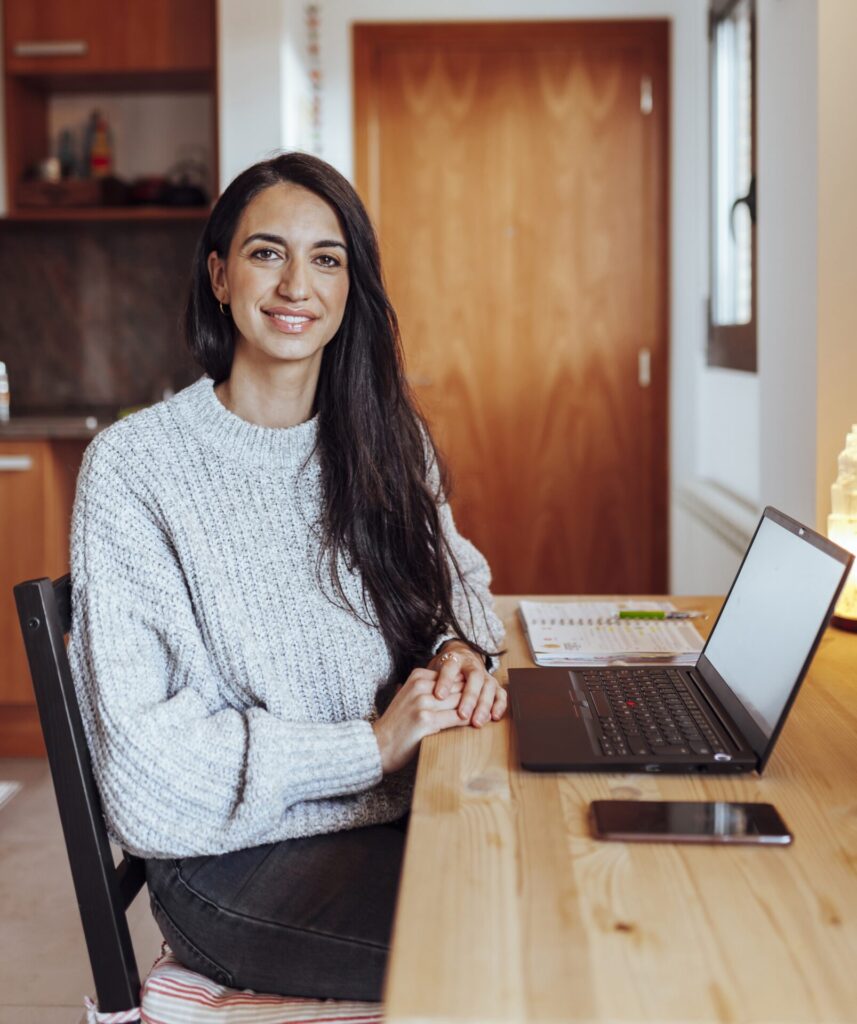 karriere tutor® Eine Frau im grauen Pullover sitzt mit Laptop, Telefon und Notizbuch an einem Tisch und lächelt, während sie sich in ihrem gemütlichen Home-Office in ihr Weiterbildungsstudium zum Wirtschaftsfachwirt vertieft.